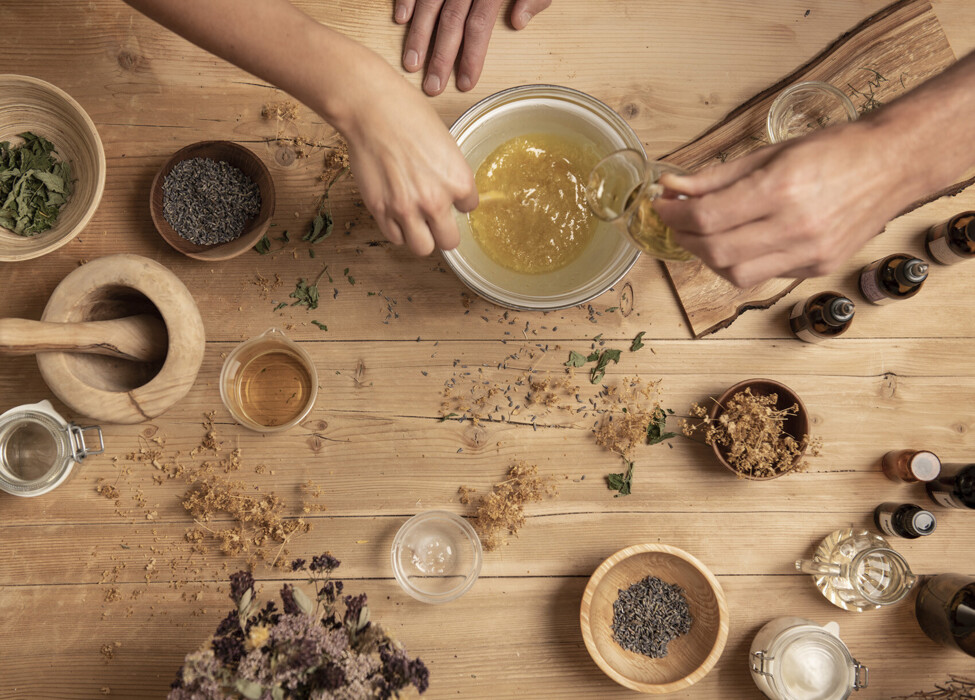 Hands preparing herbal mixture with dried ingredients on a wooden table, surrounded by jars and bowls.