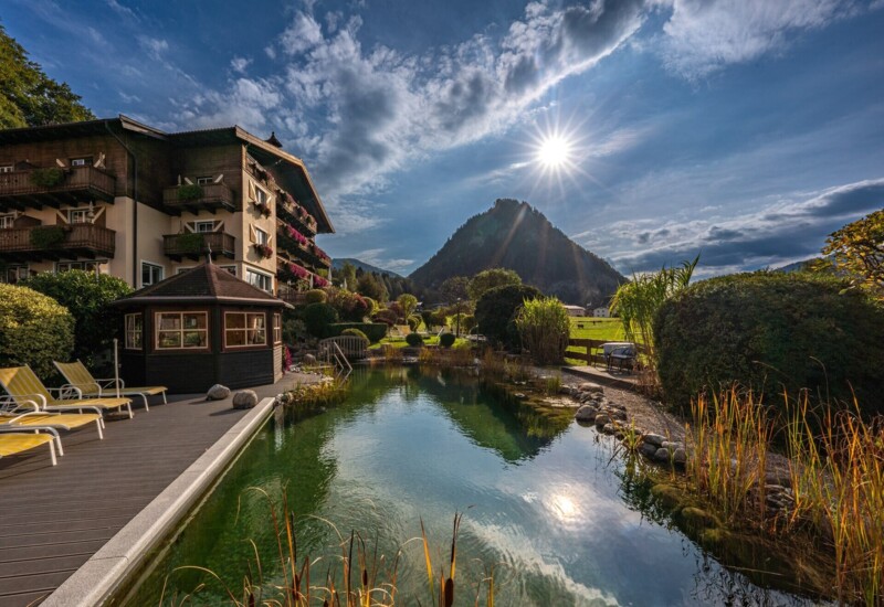 Naturbadeteich des Waldhof Fuschlsee Resorts mit Liegestühlen, umgeben von üppigem Grün, mit Bergblick und strahlendem Sonnenschein.