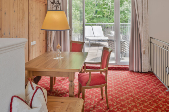 Hotel room with wooden table and red chairs, overlooking a lush balcony with sun loungers through glass doors.