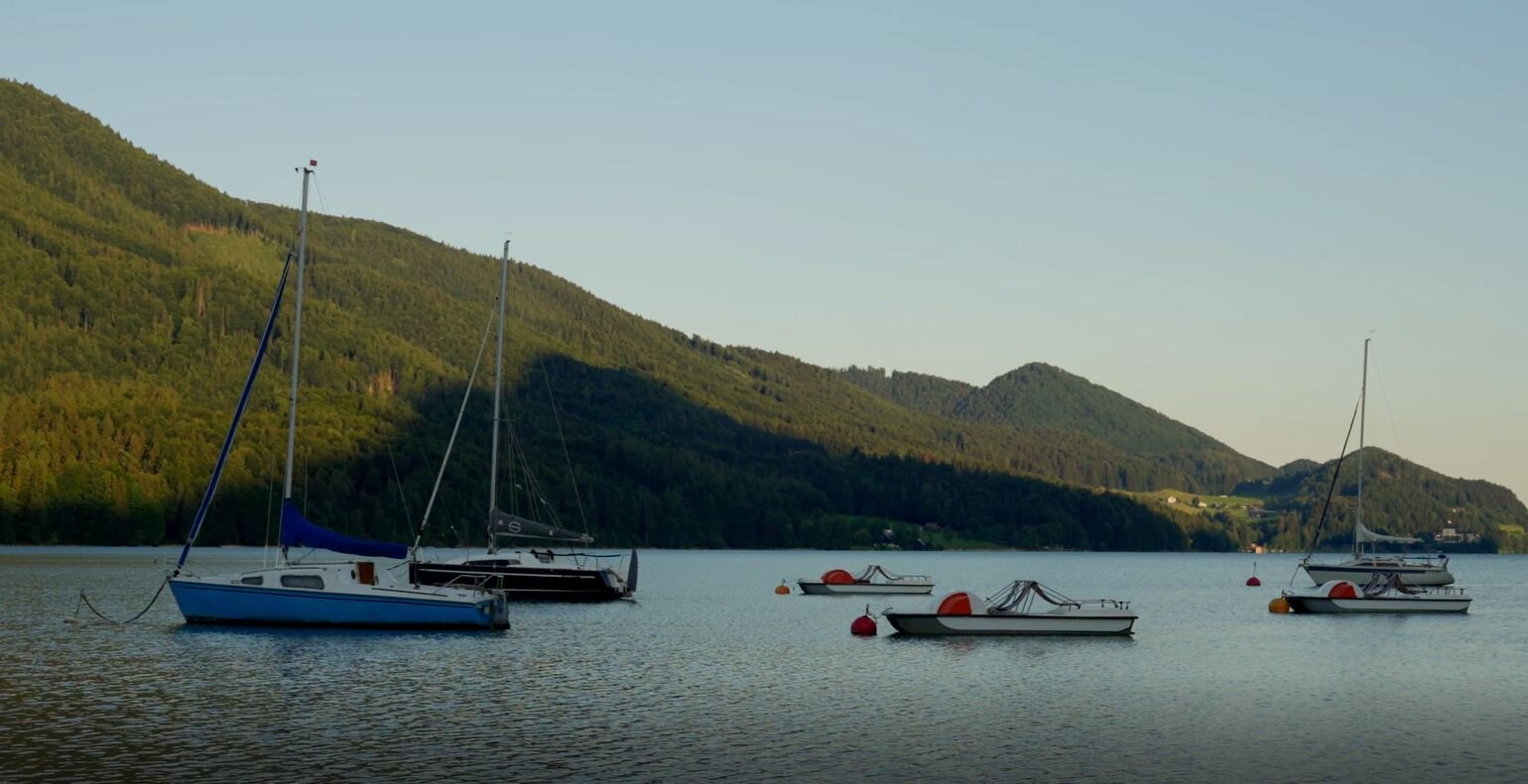 Segelboote auf dem Fuschlsee vor einer bewaldeten Bergkulisse bei Sonnenuntergang.