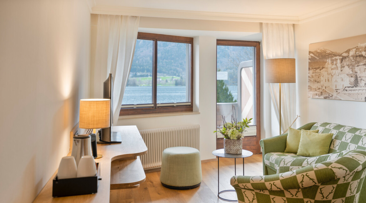 Cozy hotel room with a patterned sofa, a table with flowers, and a scenic lake view through large windows.