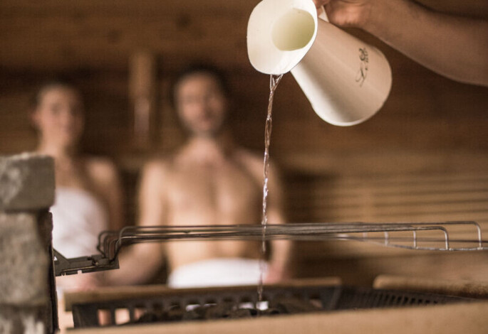 Water being poured onto sauna stones, creating steam, with two people in towels relaxing in the background.