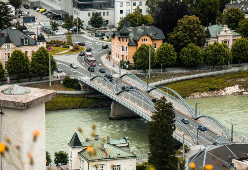 Aerial view of a scenic town with a bridge over a river, surrounded by lush greenery and historic buildings.