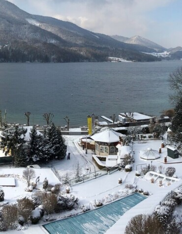 Snow-covered landscape with a lake and mountains in the background, surrounded by buildings and trees under a partly cloudy sky.