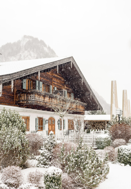 Snow-covered chalet with wooden balcony and garden, set against a mountainous backdrop.