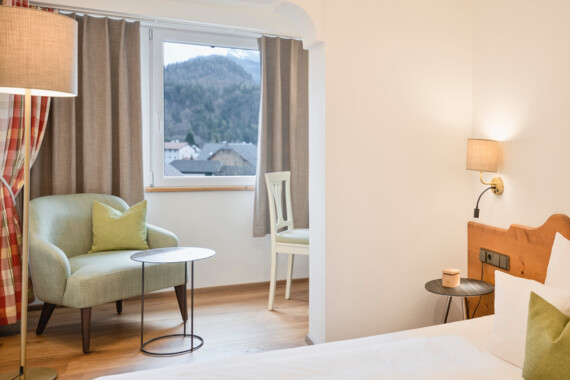 Bright hotel room with armchair, round tables, and a window view of mountains. Cozy ambiance with wooden accents and soft lighting.