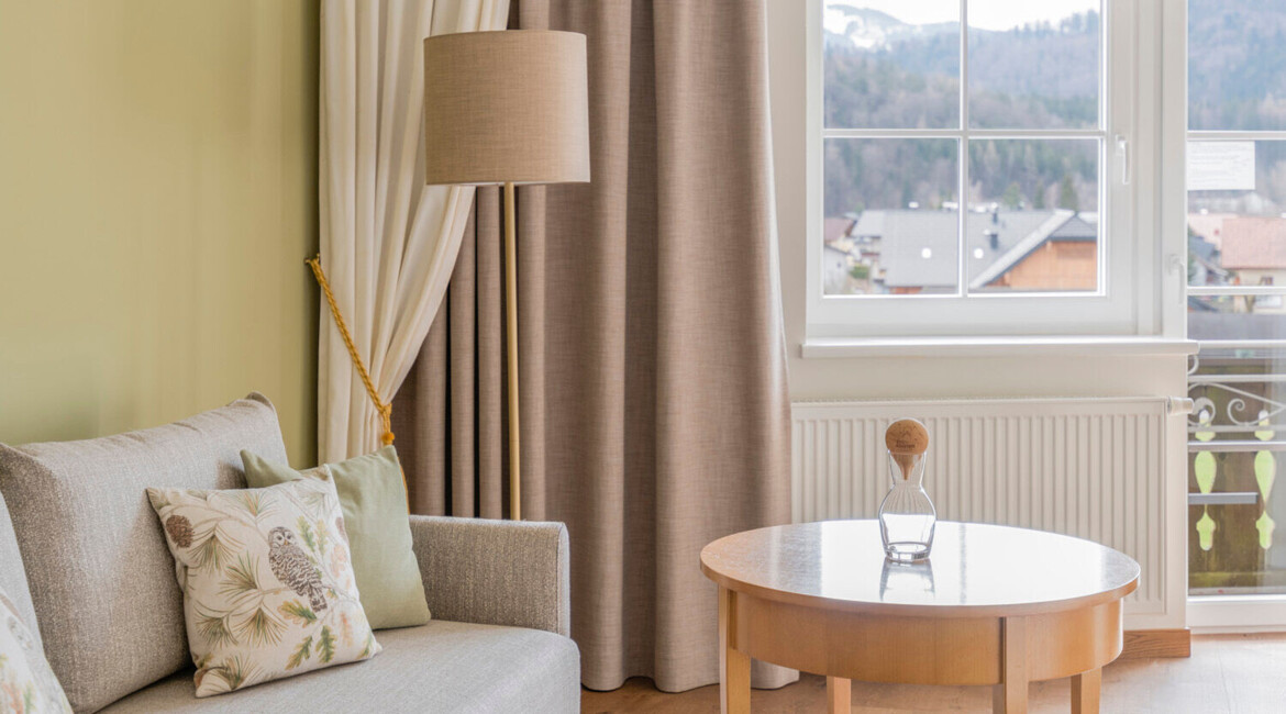 A cozy living room with a sofa, armchair, and round table, overlooking a scenic view through a window.