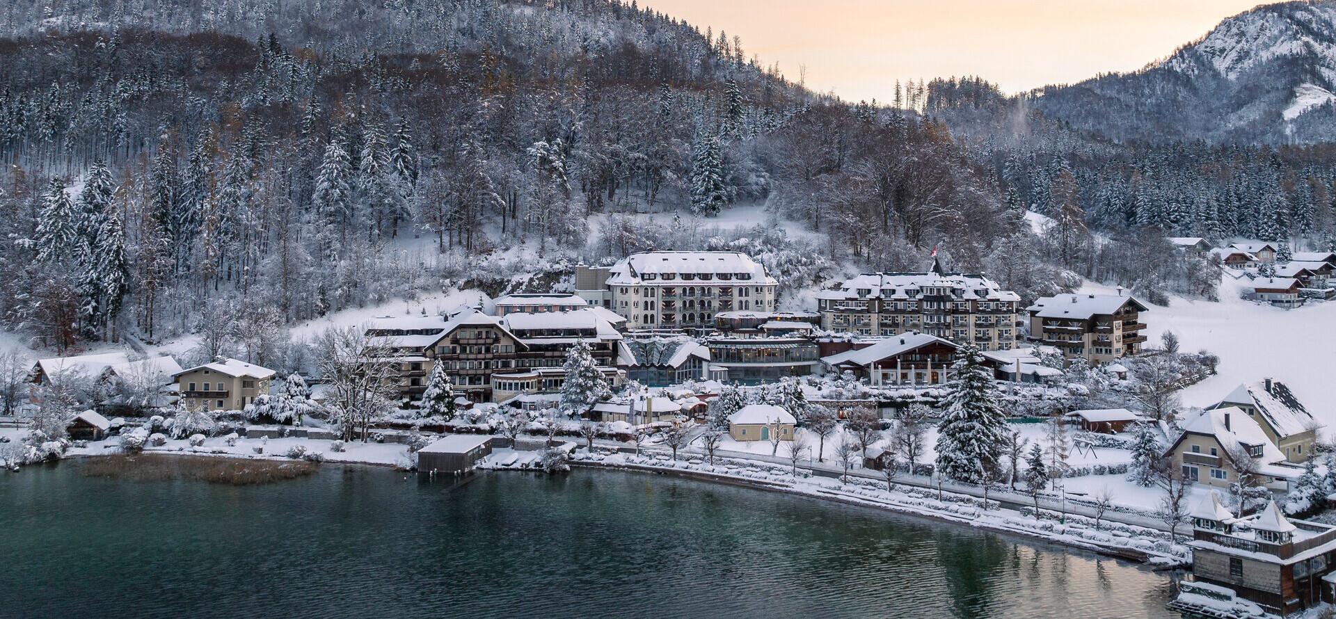 Snow-covered village by a lake with forested mountains in the background at sunrise.