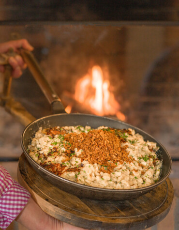 A person in a pink checkered shirt holds a pan of creamy pasta topped with crispy onions, in front of a warm fireplace.