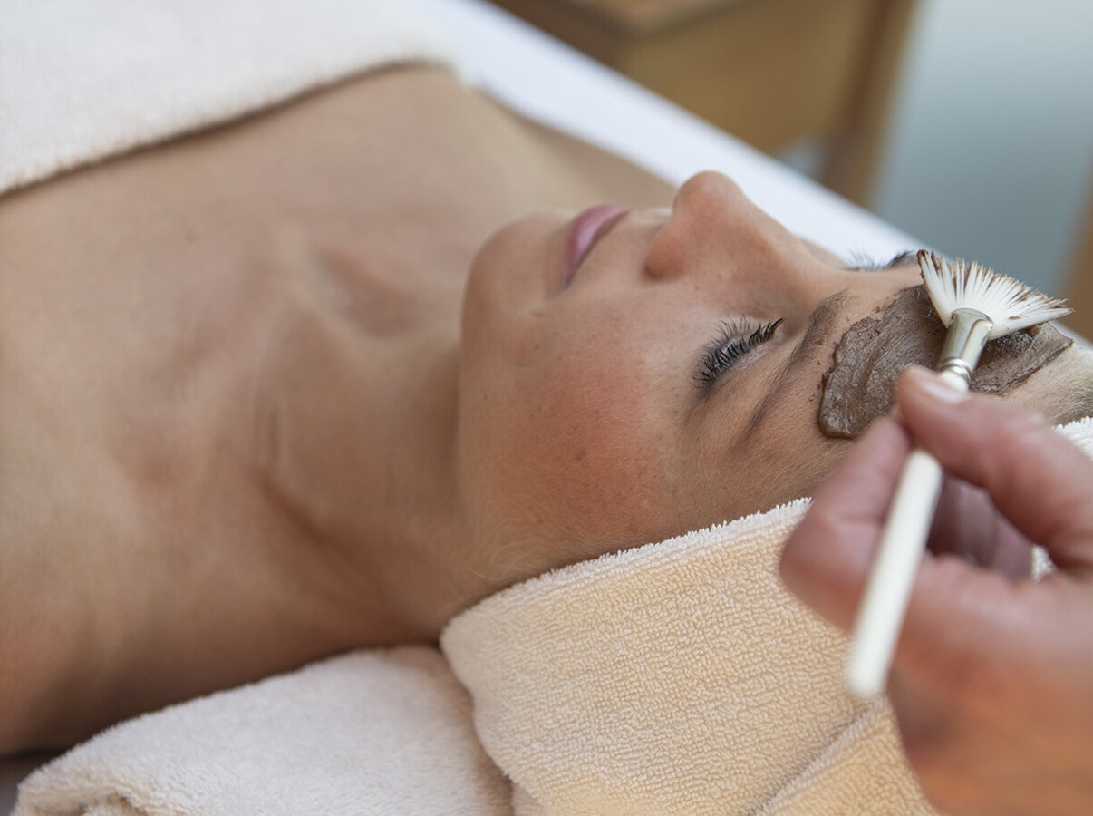 A person receiving a relaxing facial treatment with a brush, surrounded by soft towels and candles.