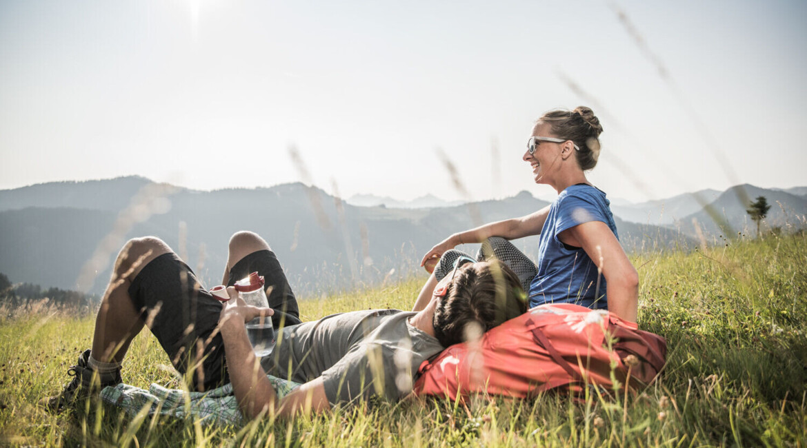 Ein Paar entspannt auf einer Bergwiese unter der Sonne, mit Blick auf die Alpen im Hintergrund.