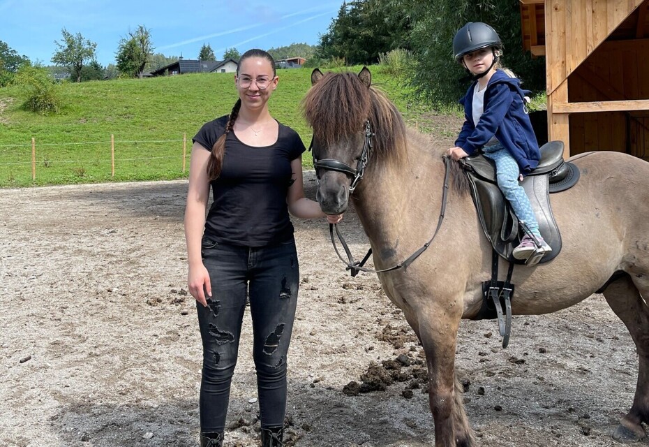 A woman stands beside a horse with a child riding it, set in a scenic outdoor area with lush greenery under a clear blue sky.