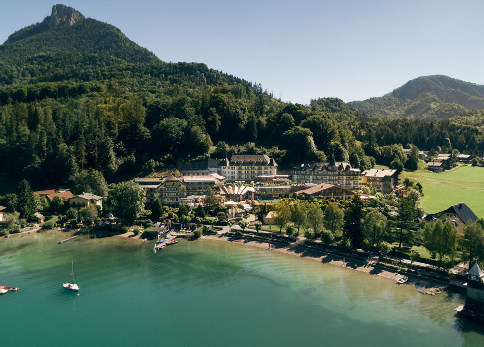 Aerial view of the Waldhof Fuschlsee Resort by a tranquil lake, surrounded by forested hills and mountains.