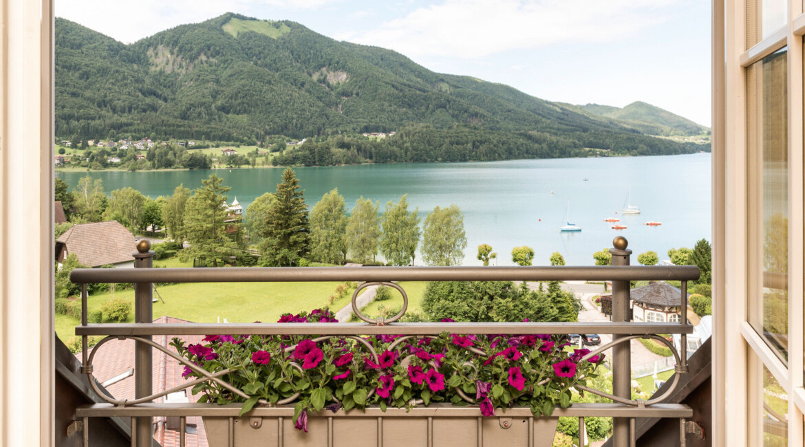 Blick von einem Balkon mit Blumen auf den malerischen Fuschlsee und die umliegenden grünen Berge.