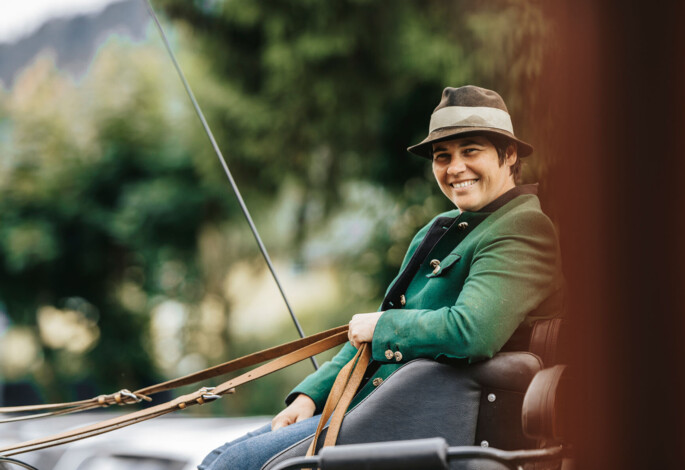 A person in a green jacket and hat smiles while sitting in a carriage, surrounded by a lush, scenic background.