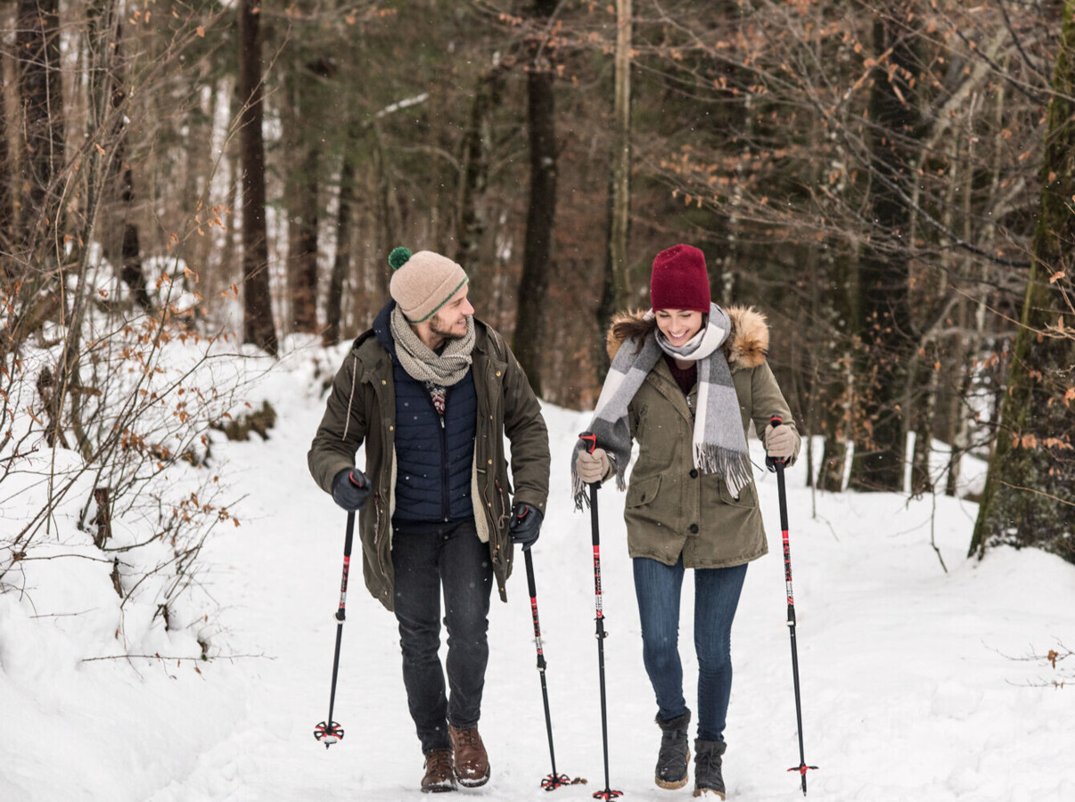 A couple enjoys a winter hike through a snowy forest, using trekking poles for support.