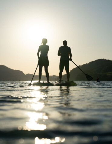 Silhouettes of two people paddleboarding on a sunlit lake with hills in the background.