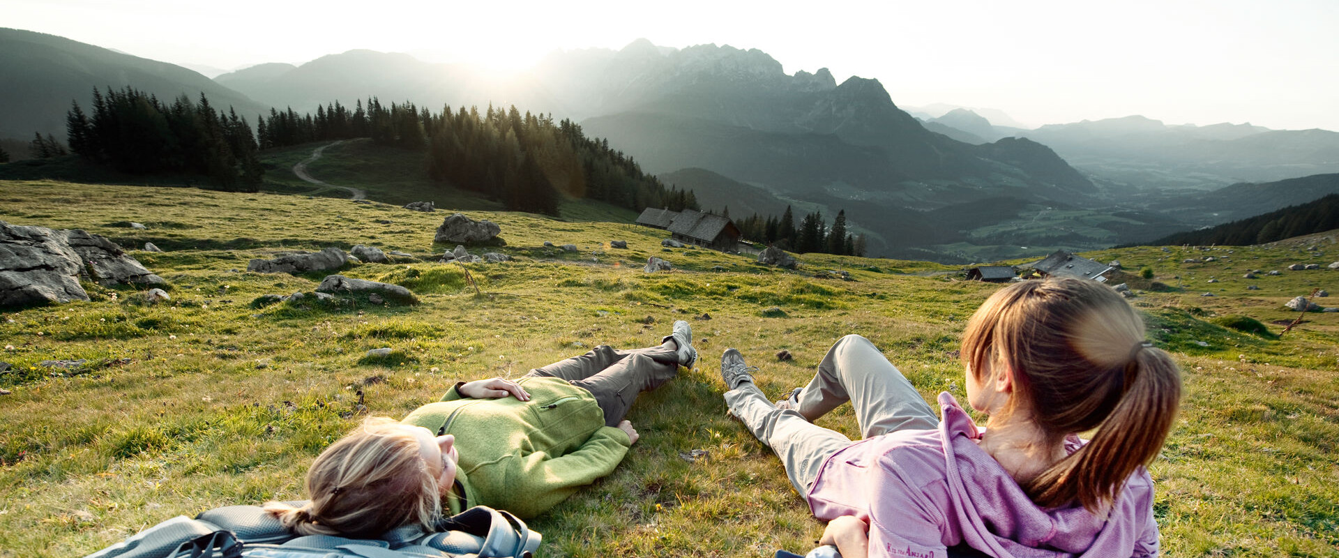 Two people relax on a grassy hillside with backpacks, enjoying a scenic mountain view at sunset.