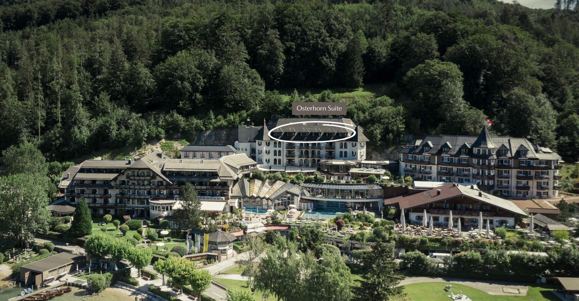 Aerial view of Waldhof Fuschlsee Resort with the Osterhorn Suite highlighted, surrounded by trees and near a lake.