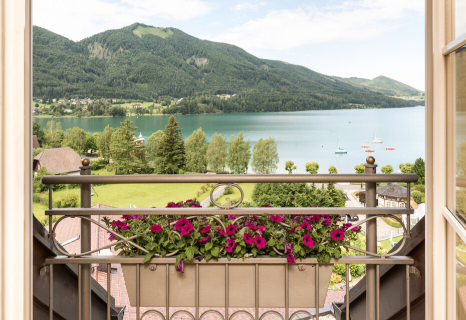View from a balcony with purple flowers, overlooking lake Fuschl and lush green mountains in the distance.