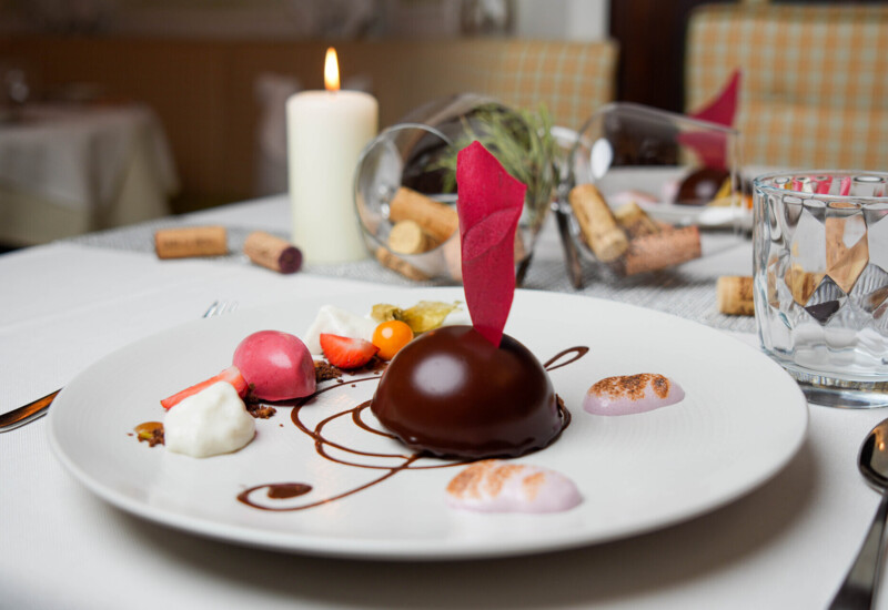 Ein kunstvoll dekoriertes Dessert auf einem weißen Teller, neben einem brennenden Kerzenlicht, auf einem elegant gedeckten Tisch.