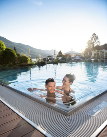 A couple enjoying a swim in an outdoor pool at Waldhof Fuschlsee Resort, surrounded by mountains and greenery.