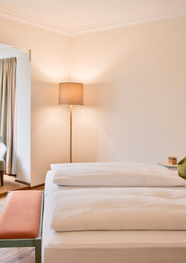 Hotel room 'Plomberg' with a cozy double bed, green pillows, seating area, and large windows overlooking trees at Waldhof Fuschlsee Resort.