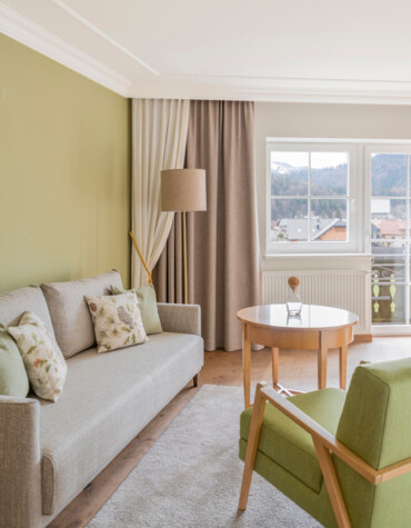 Bright hotel room with green accents, cozy sofa, armchair, and wooden table by a window offering scenic views.