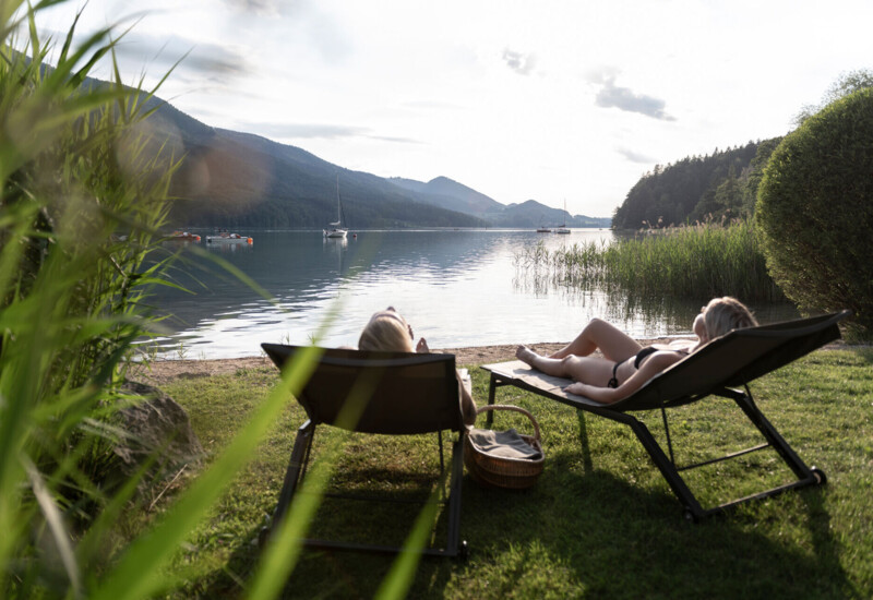 Two people relax on loungers by lake Fuschl, surrounded by lush greenery and scenic mountains in the background.