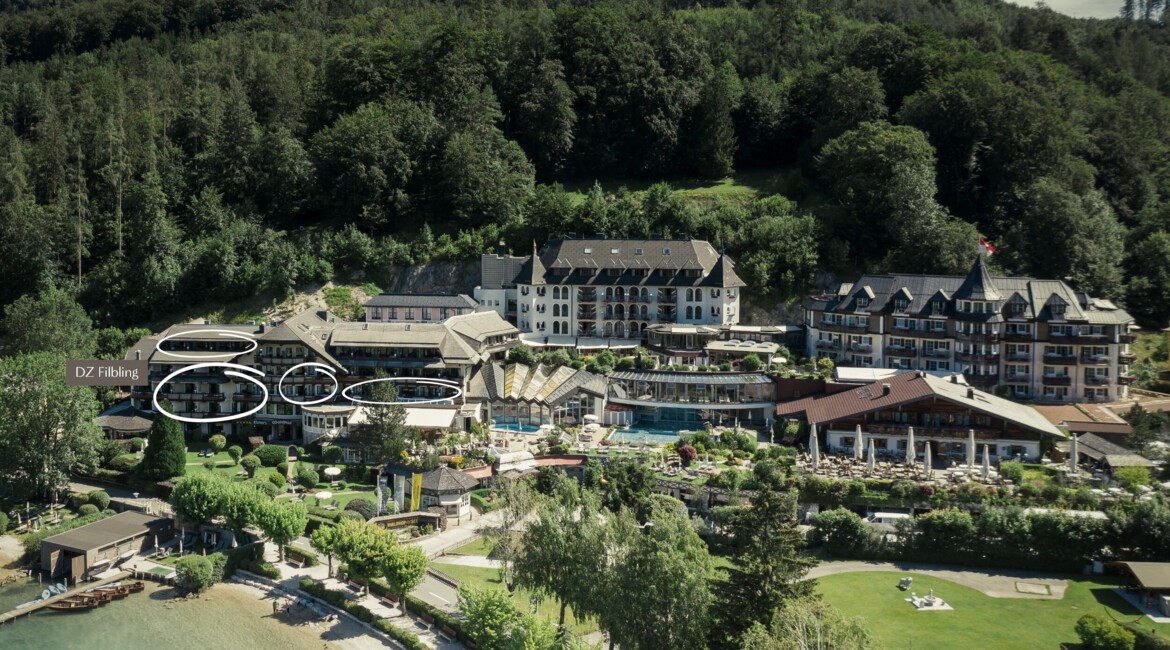 Aerial view of a scenic hotel surrounded by lush greenery and a beach, nestled against a forested hillside.