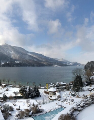 Snow-covered landscape with a lake and mountains in the background, surrounded by buildings and trees under a partly cloudy sky.