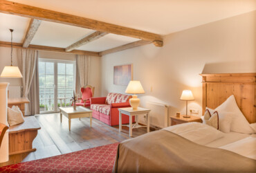 Cozy hotel room with wooden beams, a double bed, red sofa, and balcony doors. Warm lighting creates an inviting atmosphere.