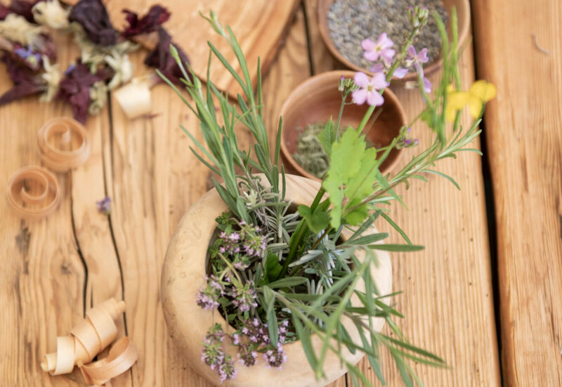 Herbs and flowers in a wooden bowl on a rustic table, surrounded by dried flowers, wooden shavings, and a pestle.