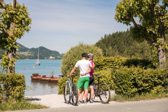 Ein Paar mit Fahrrädern genießt die Aussicht auf den Fuschlsee, umgeben von grüner Landschaft und klarem Himmel.