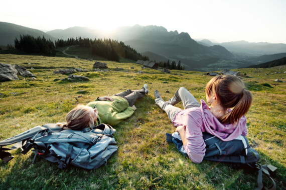 Two people relax on a grassy hillside with backpacks, enjoying a scenic mountain view at sunset.