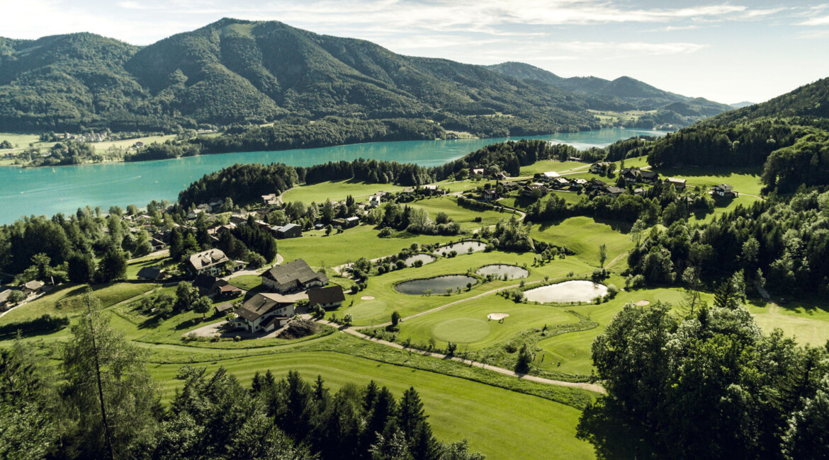 Scenic aerial view of a lush golf course surrounded by mountains and a lake.