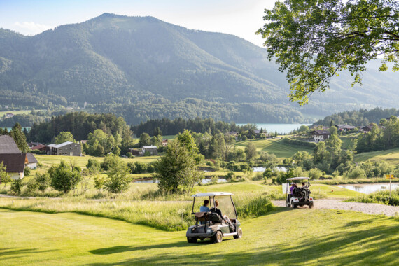 Zwei Golfcarts fahren über einen grünen Golfplatz mit Blick auf Berge und den Fuschlsee bei sonnigem Wetter.