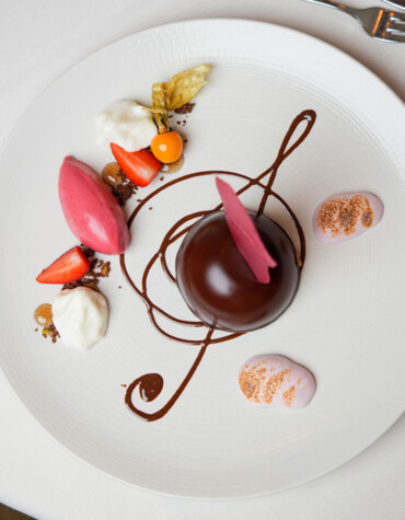Elegant dessert with chocolate dome, strawberry sorbet, cream, and decorative chocolate swirl on a white plate.