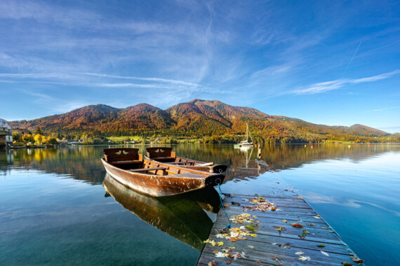 Ein Holzboot liegt an einem Steg auf einem ruhigen See, umgeben von herbstlichen Bergen unter klarem blauem Himmel.