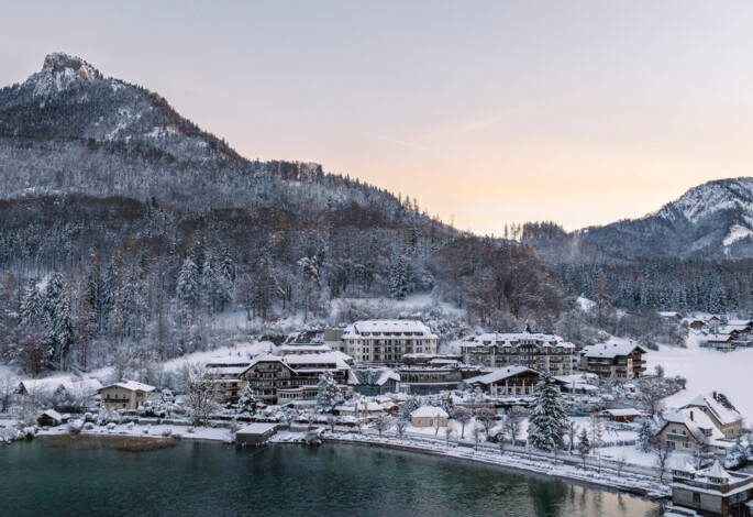 Snow-covered village by a lake with forested mountains in the background at sunrise.