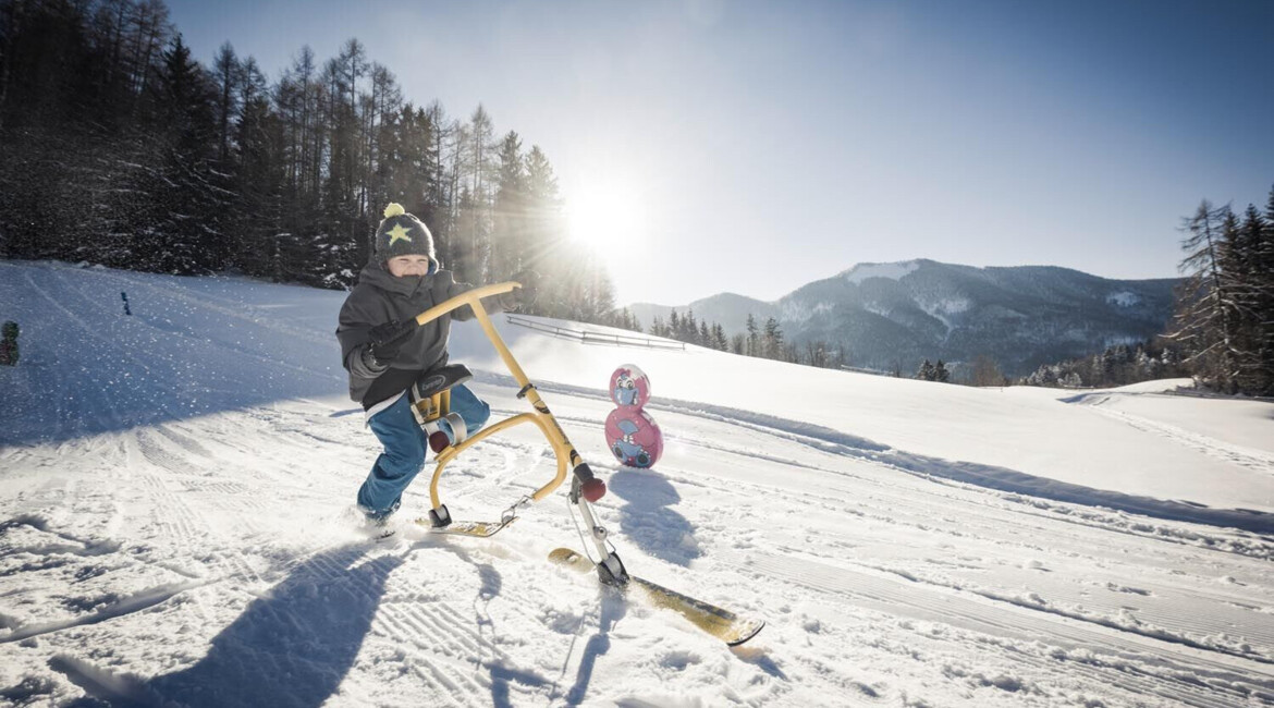 Child sledding on a snowy hill with a scenic mountain backdrop under a clear blue sky.