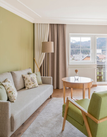 Bright living room with green accents, cozy sofa, armchair, and wooden table by a window offering scenic views.