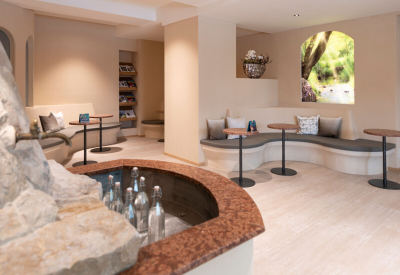 Cozy seating area with curved benches and small tables, featuring a scenic window view of lush greenery and a stone fountain with water bottles.