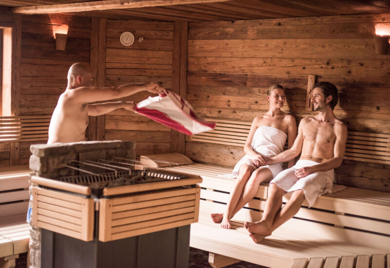 People enjoying a relaxing sauna session, with one person creating steam by pouring water on hot stones.