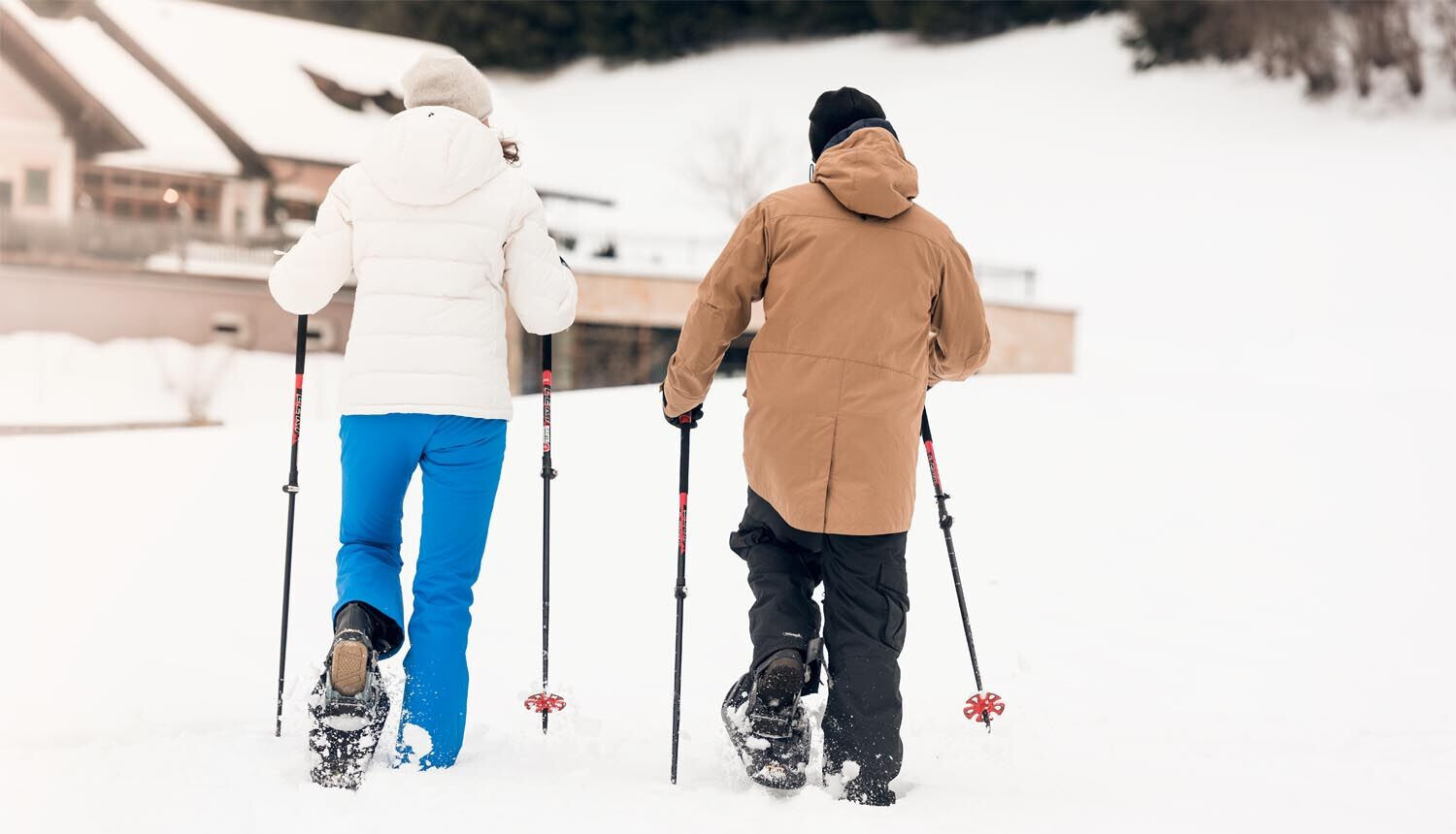 Two people snowshoeing on a snowy landscape, wearing winter gear and using poles, with a building in the background.