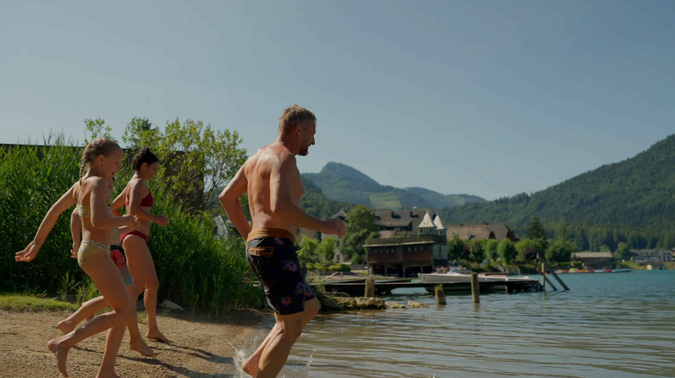Family enjoying the beach at Waldhof Fuschlsee Resort, with mountains in the background under a clear sky.