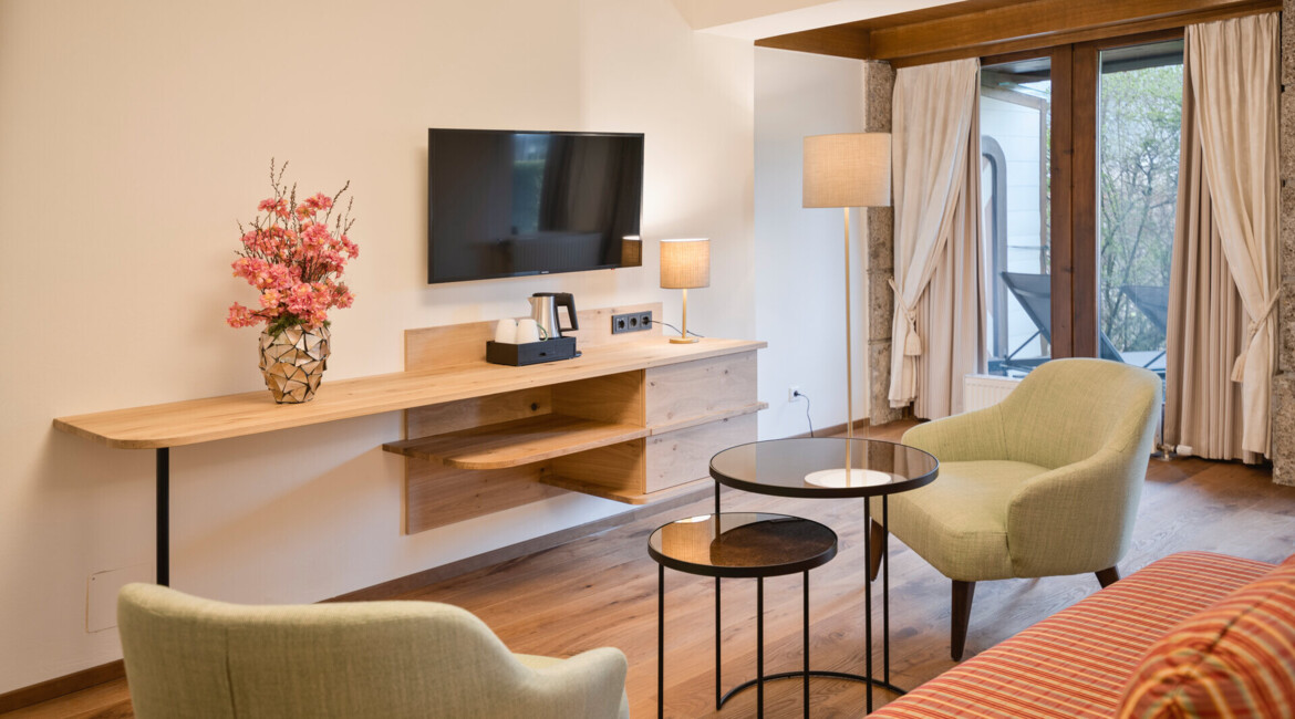 Modern hotel room with a TV, elegant seating, and a vase of pink flowers. Large window with curtains brings natural light into the space.