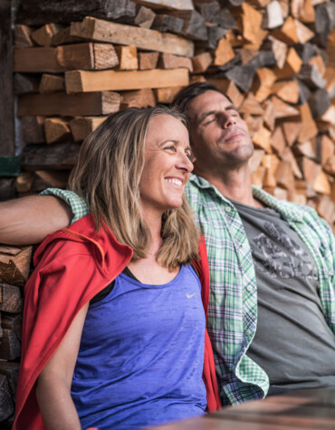A couple relaxes contentedly in front of a house wall and enjoys a peaceful moment, with stacked firewood in the background.