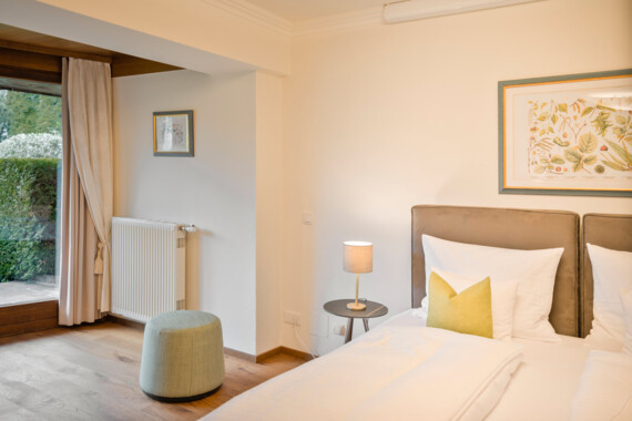 Hotel room with a bed, side table, and lamp, featuring garden views through a large window.