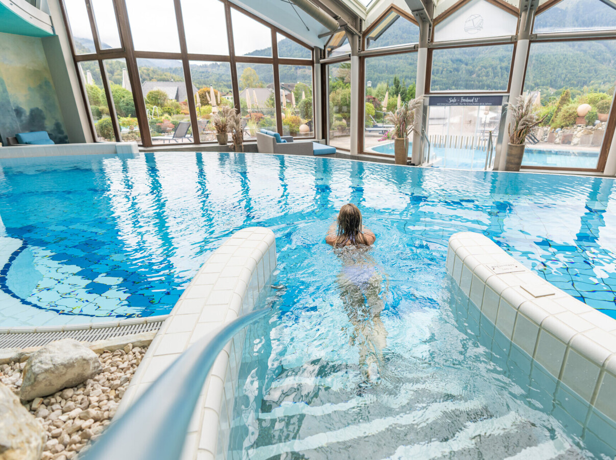 A person swims in an indoor pool with large windows at Waldhof Fuschlsee Resort, offering views of the surrounding landscape.